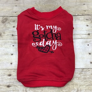 May include: Red dog shirt featuring the text "It's my Gotcha Day" in white and black script. A black heart with a dotted pattern and an arrow is below the text. The shirt is made of a soft material.