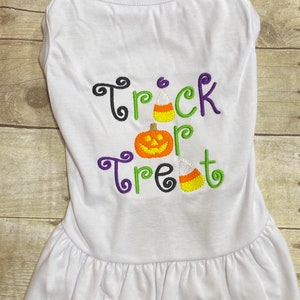 K&ouml;nnte beinhalten: Wei&szlig;es Hundekleid mit R&uuml;schensaum. Auf der Vorderseite des Kleides sind die gestickten Worte "Trick or Treat" in Schwarz, Gr&uuml;n und Lila sowie ein K&uuml;rbis und Zuckermais abgebildet.