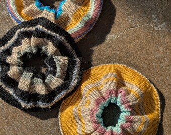 May include: Three knitted hair scrunchies in different color combinations. The scrunchies are yellow, beige, black, and pink with a blue stripe.