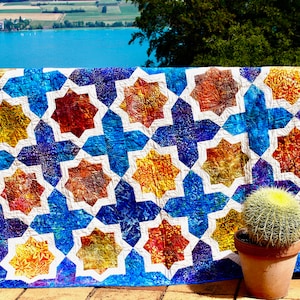 Puede incluir: Una colcha colorida con un patrón geométrico de estrellas en tonos de azul, blanco, naranja y rojo. La colcha está colgada en una barandilla con un cactus en una maceta en primer plano.