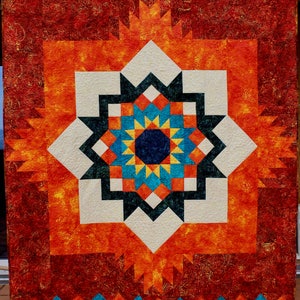May include: A quilt with a geometric pattern in shades of orange, red, white, blue, and yellow. The center of the quilt features a starburst design.