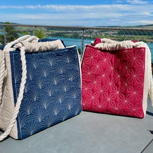 May include: Two tote bags with rope handles. One bag is navy blue with a white fan pattern, the other is red with a white star pattern. Both bags have a beige drawstring closure and are made of quilted fabric.