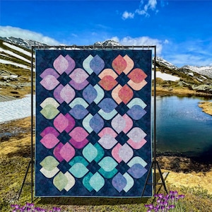 Puede incluir: Una colcha con un patrón geométrico de formas de lágrima superpuestas en tonos de rosa, azul, verde y turquesa sobre un fondo azul oscuro. La colcha se muestra en un soporte frente a un lago de montaña.