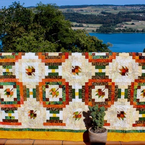May include: A quilt with a patchwork design featuring a variety of fall colors, including orange, brown, green, and yellow. The quilt is made up of squares and rectangles, and each square features a different pattern. The quilt is hanging on a fence against a backdrop of a lake and trees.