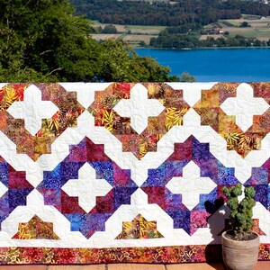 May include: A quilt with a white background and a pattern of colourful squares and crosses. The quilt is made of fabric with a batik design. The quilt is hanging on a fence in front of a lake and trees.