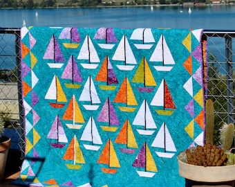 May include: A turquoise quilt with a repeating pattern of sailboats. The boats have white sails and hulls in shades of orange, yellow, purple, and red. The quilt is shown outdoors, with a lake and hills in the background.