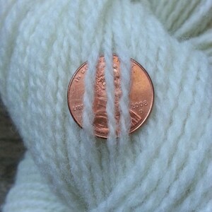 May include: A close-up of a ball of white yarn with a copper-colored penny resting on top. The penny is dated 2002 and has the words "IN GOD WE TRUST" and "LIBERTY" visible.