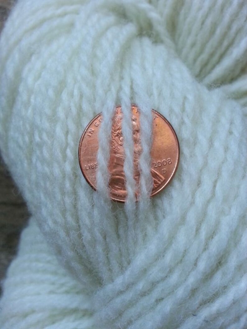May include: A close-up of a ball of white yarn with a copper-colored penny resting on top. The penny is dated 2002 and has the words "IN GOD WE TRUST" and "LIBERTY" visible.