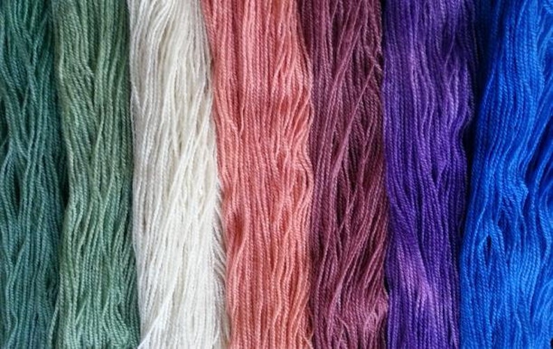 May include: Close-up of seven skeins of yarn in various colors, including green, white, pink, purple, blue, and a darker blue. The yarn is arranged vertically, with each skein showing its texture and color.