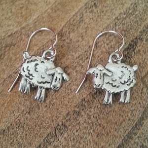 May include: Silver dangle earrings featuring a cute sheep design. The sheep are silver with a textured wool pattern.