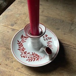 Può includere: Portacandele in ceramica bianca con un motivo floreale rosso e un manico rosso. Una candela rossa è nel portacandele.