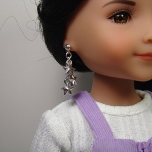 May include: A doll with black hair wearing a white shirt and purple overalls. The doll is wearing silver star-shaped dangle earrings.