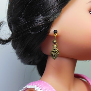 May include: A close-up of a doll wearing a gold-colored leaf-shaped earring. The earring has a black bead at the top and a small, round bead in the middle. The doll has black hair and is wearing a pink top.