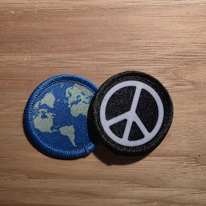 May include: Two circular embroidered patches. One patch features a blue and light green map of the Earth. The second patch is black with a white peace symbol centered within a circular border. Both patches have a stitched edge finish.