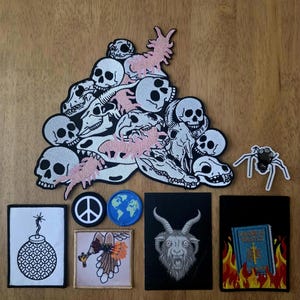 Pode incluir: Uma coleção de patches bordados incluindo uma grande pilha de crânios brancos e centopeias rosas, uma aranha preta, uma bomba branca, um símbolo da paz, um globo azul, um pássaro, uma cabeça de cabra e um livro azul em chamas.