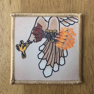 May include: A square embroidered patch featuring a graphic illustration of a bird of prey with tan and white feathers. The bird holds a bundle of arrows with red fletching in its yellow talons, with a bright orange flame effect at the arrow tips.