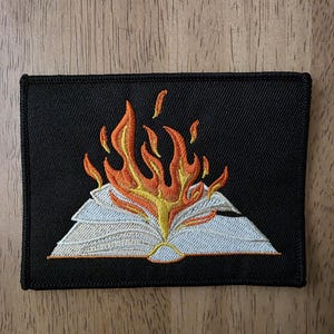 May include: A rectangular black embroidered patch featuring a design of an open book with white pages. Bright orange and yellow flames rise from the center of the book, with a thick black stitched border framing the entire rectangular shape.