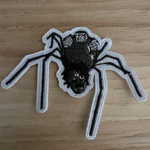Op de afbeelding: Een geborduurde strijkpatch in de vorm van een spin. De spin heeft een zwart lichaam met witte cirkelvormige markeringen op de rug en lange, dunne zwarte poten. Het hele ontwerp is afgewerkt met een witte rand voor een opvallend contrast.