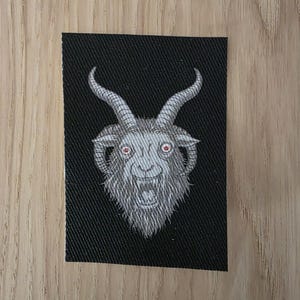May include: A rectangular black fabric patch featuring an embroidered illustration of a goat head. The goat is rendered in shades of grey and white with prominent curved horns and small, distinct red eyes, showing an open mouth with visible teeth.