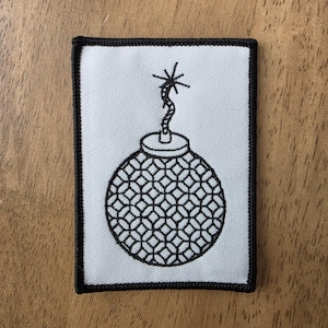 May include: A rectangular embroidered patch featuring a white background with a black border. The design depicts a round bomb with a textured, geometric pattern on its surface and a lit fuse with a star-shaped spark at the top in black thread.