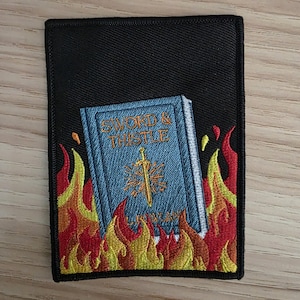 May include: A rectangular black embroidered patch featuring a light blue book with the text SWORD & THISTLE and a sword icon on the cover. The book is surrounded by vibrant yellow, orange, and red flames embroidered against the black background.