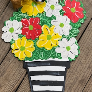 May include: A wooden door hanger shaped like a flower pot with a black and white striped pattern. The pot is filled with yellow, red, and white flowers with green leaves.