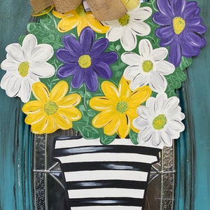 May include: A wooden door wreath featuring a bouquet of yellow, white, and purple flowers in a black and white striped pot. The flowers are painted in a realistic style and the pot has a simple design.