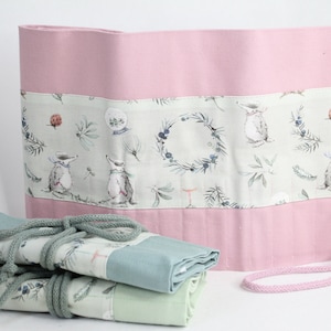 Könnte beinhalten: Eine rosa Stoff-Bleistiftrolle mit einer grünen und weißen Stoffauskleidung, die ein Muster aus kleinen Tieren und Grünpflanzen zeigt. Die Rolle ist aufgerollt und mit einer grünen Schnur gebunden.
