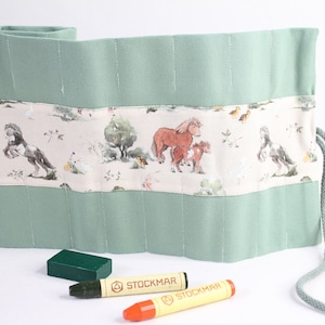 Könnte beinhalten: Eine grüne Stoff-Bleistiftrolle mit einem Pferde- und Bauernhoftier-Druck. Die Rolle hat einen Kordelverschluss und ist ausgerollt, um die einzelnen Fächer für Bleistifte zu zeigen. Zwei Buntstifte liegen vor der Rolle.