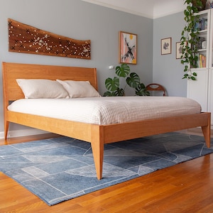 May include: A wooden bed frame with a light wood finish and a white bedspread. The bed is sitting on a blue and white patterned rug. There is a plant in a pot on the left side of the image.