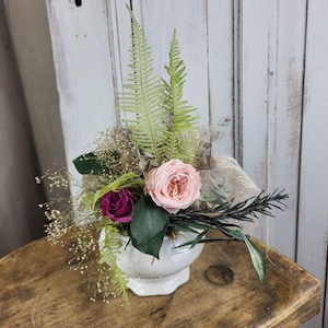 Könnte beinhalten: Ein Blumenarrangement in einer weißen Keramikvase mit getrockneten Blumen und Grünzeug. Es beinhaltet eine rosa Rose, eine tiefviolette Rose sowie verschiedene Farne und Blätter. Die Vase steht auf einer Holzoberfläche und erzeugt eine rustikale Ästhetik.