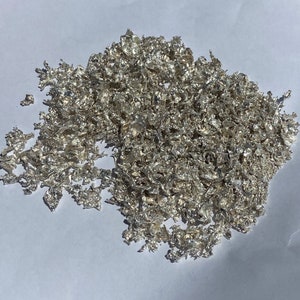 May include: A pile of silver flakes, resembling a cluster of small, irregular, metallic leaves. The flakes are a silvery-white color and have a slightly textured surface.
