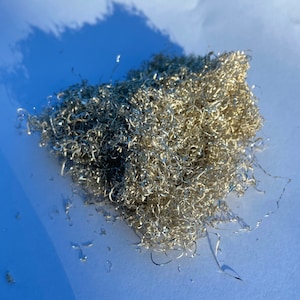 May include: A collection of silver metal shavings, likely from machining, sits on a white surface. The tightly curled shavings reflect light, giving a bright, metallic look. The image is captured in natural light, with shadows visible.