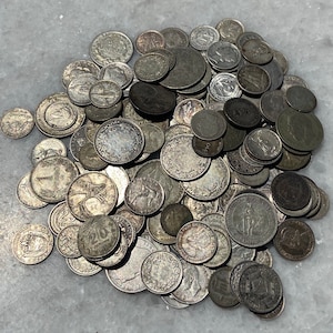 May include: A large collection of silver coins, many with intricate designs, are scattered on a white surface. The coins are of various sizes and shapes, with some featuring raised edges and others with smooth surfaces.