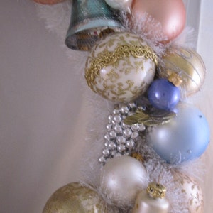 K&ouml;nnte beinhalten: Eine wei&szlig;e, goldene und blaue Weihnachtsgirlande mit Ornamenten, Glocken und Perlen. Die Girlande ist mit wei&szlig;en Federn verziert und hat einen Vintage-Look.