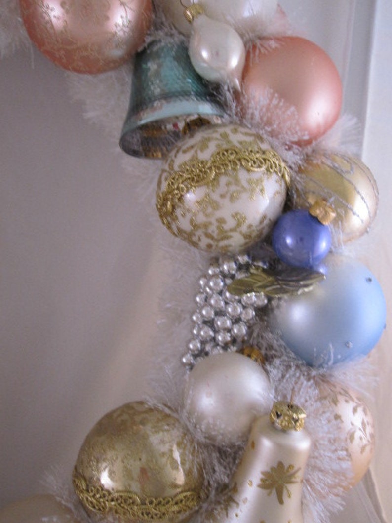 K&ouml;nnte beinhalten: Eine wei&szlig;e, goldene und blaue Weihnachtsgirlande mit Ornamenten, Glocken und Perlen. Die Girlande ist mit wei&szlig;en Federn verziert und hat einen Vintage-Look.