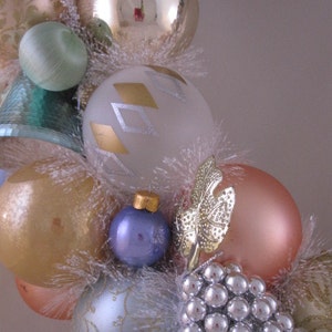 K&ouml;nnte beinhalten: Ein Weihnachtskranz, der mit verschiedenen Ornamenten geschm&uuml;ckt ist, darunter goldene, silberne und blaue Glaskugeln sowie wei&szlig;er, flauschiger Besatz. Der Kranz ist au&szlig;erdem mit einer Gruppe silberner Perlen verziert.