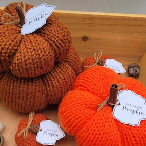 May include: A wooden crate filled with handmade knitted pumpkins. The pumpkins are in shades of orange and brown and have tags that say "Handmade Pumpkin".