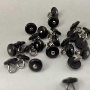 May include: A collection of small, round black buttons with a smooth finish and a metallic center detail. Each button features a silver-toned metal shank loop on the underside for attachment, arranged in a loose cluster on a plain white surface.