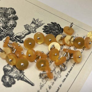 May include: A collection of small, round, translucent amber-colored buttons with a smooth, polished finish. Each button features a central silver-toned metal shank on the back for attachment, arranged in a cluster on a cream-colored paper background.