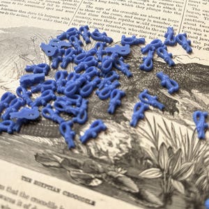 May include: A collection of small, blue, plastic crocodile-shaped game pieces scattered across a vintage book page featuring an illustration of a crocodile and text about the Egyptian crocodile, with each piece showing a detailed reptilian form.
