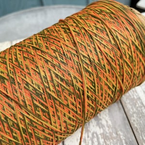 May include: A large cylindrical spool of yarn featuring a variegated color pattern of vibrant orange, deep forest green, and muted olive tones. The thread is tightly wound in a consistent diagonal cross-hatch texture across the entire surface.