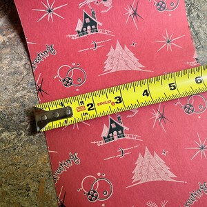 May include: A yellow tape measure with black markings is placed on top of red wrapping paper with a black and white pattern of houses, trees, stars, and the word "greetings". The tape measure reads 2 feet and 6 inches.