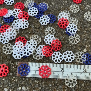 May include: A collection of red, white, and blue decorative flower-shaped embellishments scattered on a gray surface. A ruler is placed on the surface with the numbers 1, 2, 3, 4, 5, 6, 7, 8, and 9 visible.