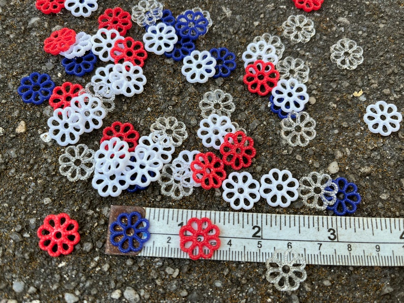 May include: A collection of red, white, and blue decorative flower-shaped embellishments scattered on a gray surface. A ruler is placed on the surface with the numbers 1, 2, 3, 4, 5, 6, 7, 8, and 9 visible.