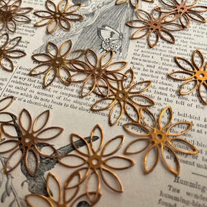 May include: A collection of 15 gold metal flower-shaped embellishments. The embellishments are delicate and intricate, with a circular center and six petals.