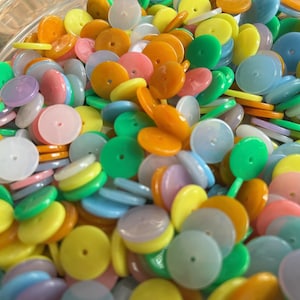 May include: A close-up shot of a clear bowl filled with a variety of colorful, flat, round plastic beads. The beads are in shades of yellow, orange, green, pink, blue, and purple. Each bead has a small hole in the center.
