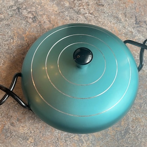 May include: A teal-colored, round cooking pot with a black knob and handles. The pot has concentric silver rings and black, looped handles on either side. The pot is sitting on a mottled brown and gray surface.