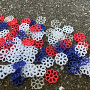 May include: A collection of red, white, and blue plastic flower-shaped embellishments. The embellishments are scattered on a gray surface.
