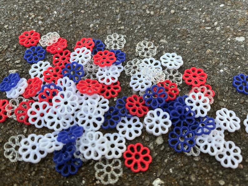 May include: A collection of red, white, and blue plastic flower-shaped embellishments. The embellishments are scattered on a gray surface.